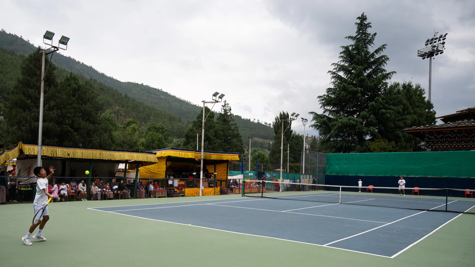 An evening match under the lights at the National Tennis Centre, spectators along the yellow canopy and the Thimphu hills behind