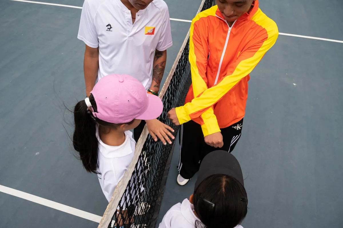A BTF coach leaning over the net to speak with a young player in a pink cap