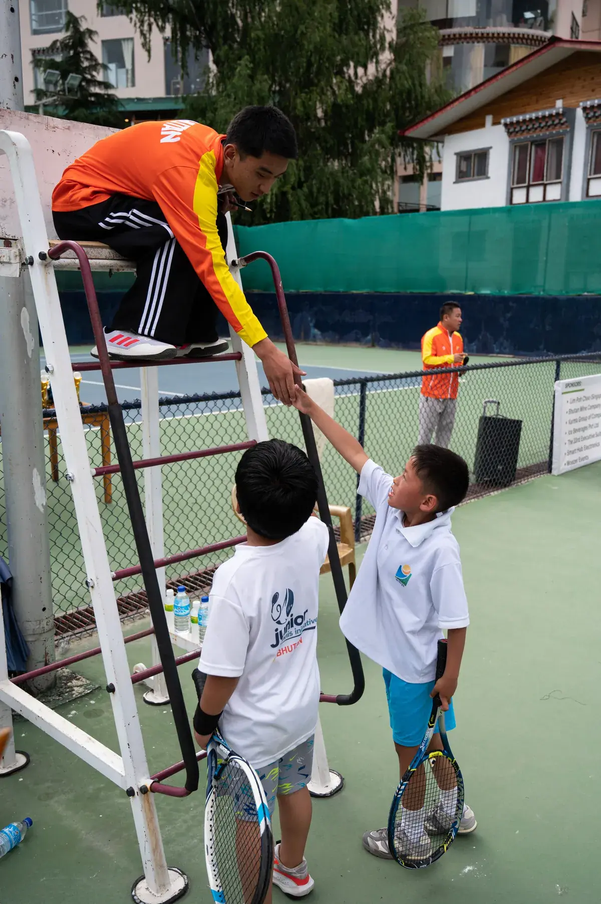 A coach on the umpire's chair reaching down to hand a racquet to two small players