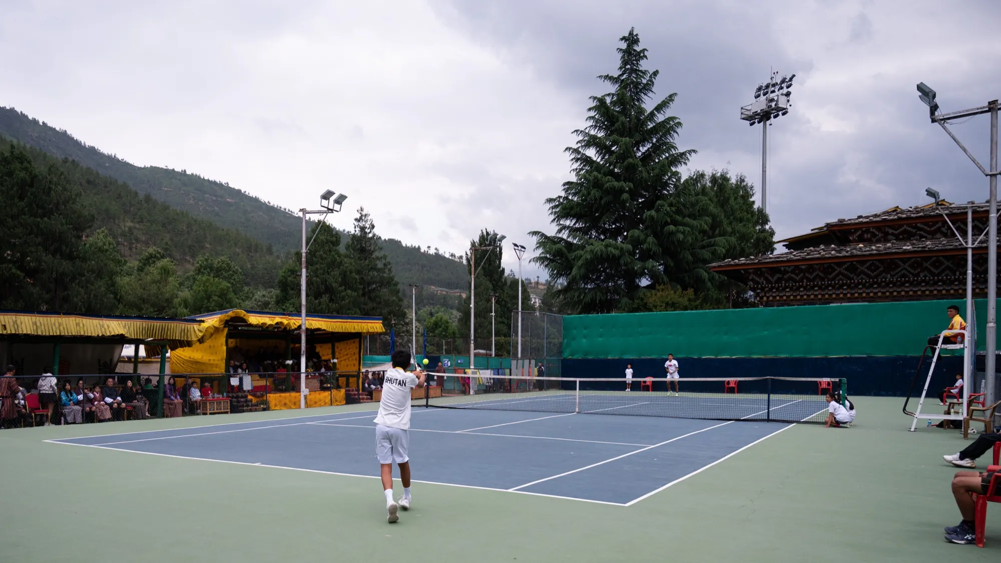 An evening doubles match at the National Tennis Centre, spectators under the yellow canopy and forested hills behind