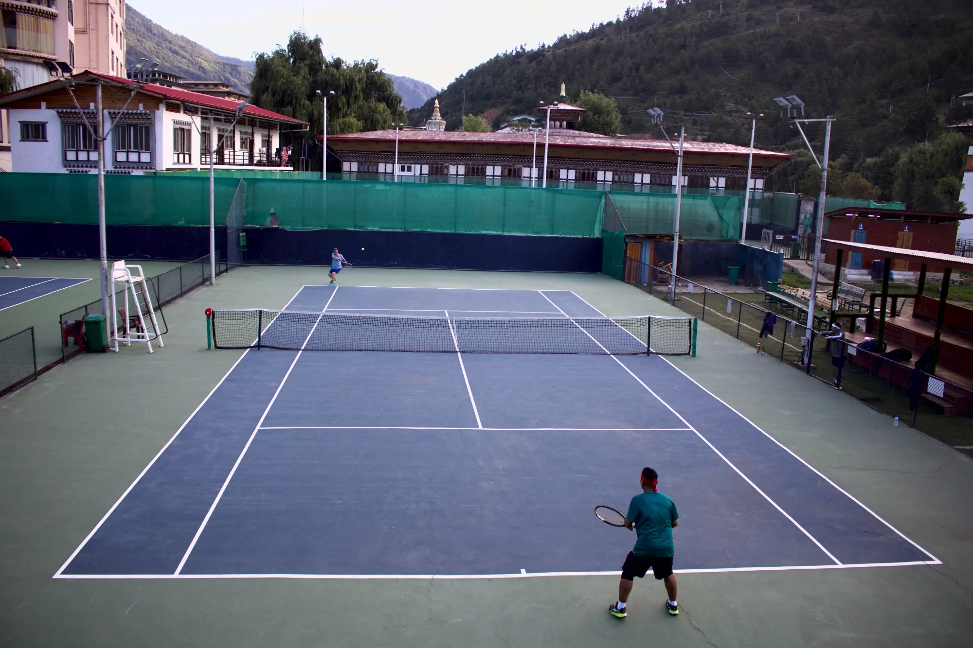 An evening singles match at the National Tennis Centre, traditional Bhutanese buildings and the forested Thimphu hills behind