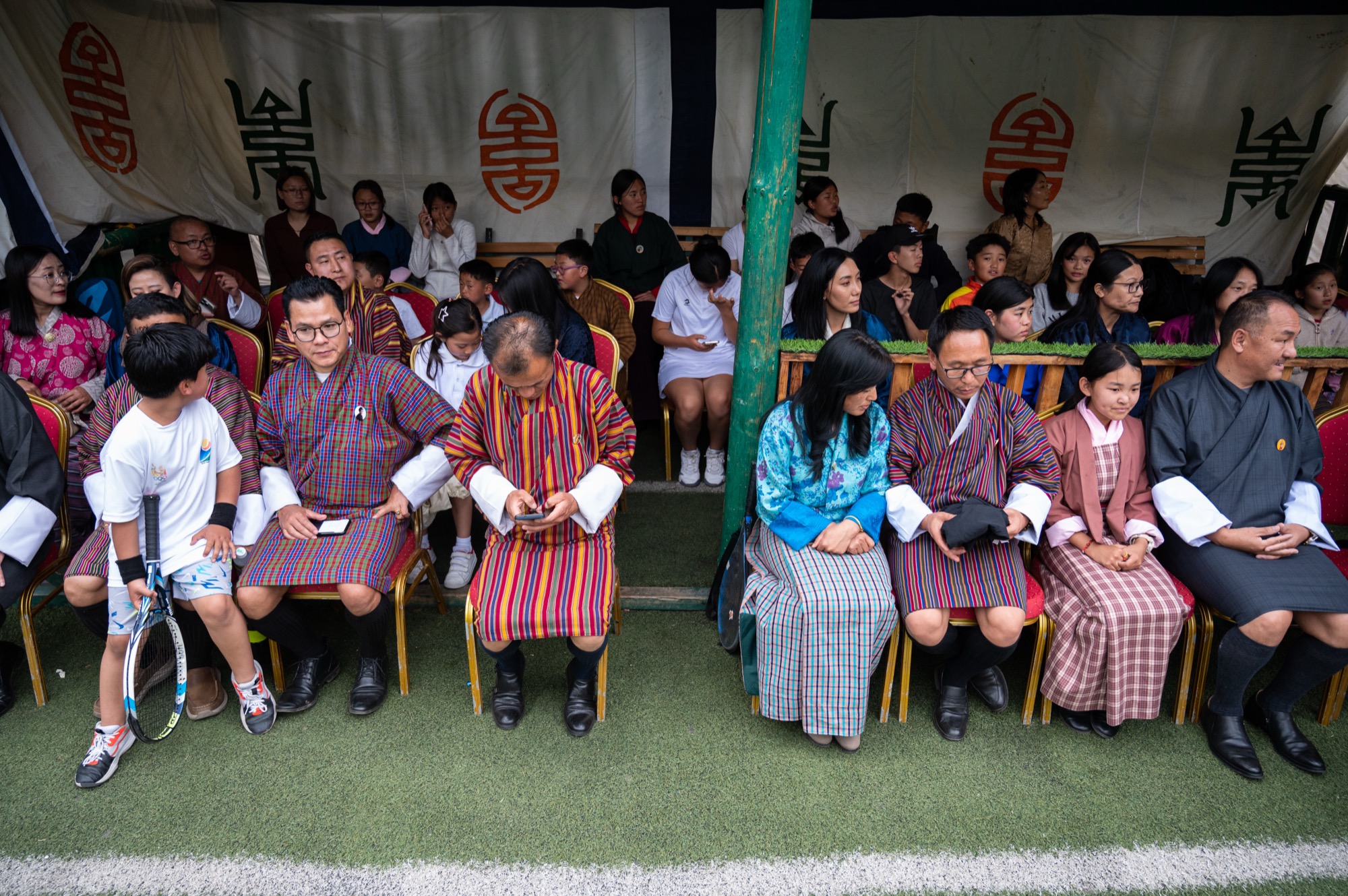 Families seated courtside under the canopy, children with racquets beside their parents