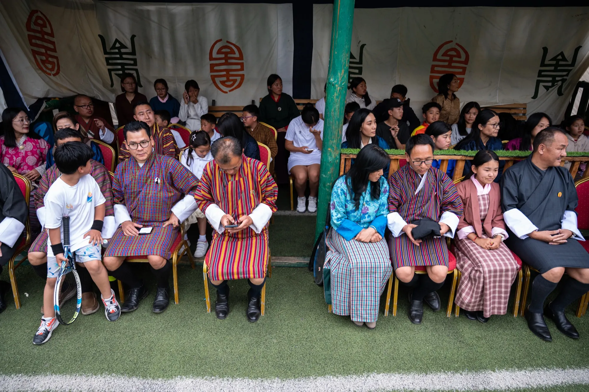 Families seated courtside under the canopy, children with racquets beside their parents