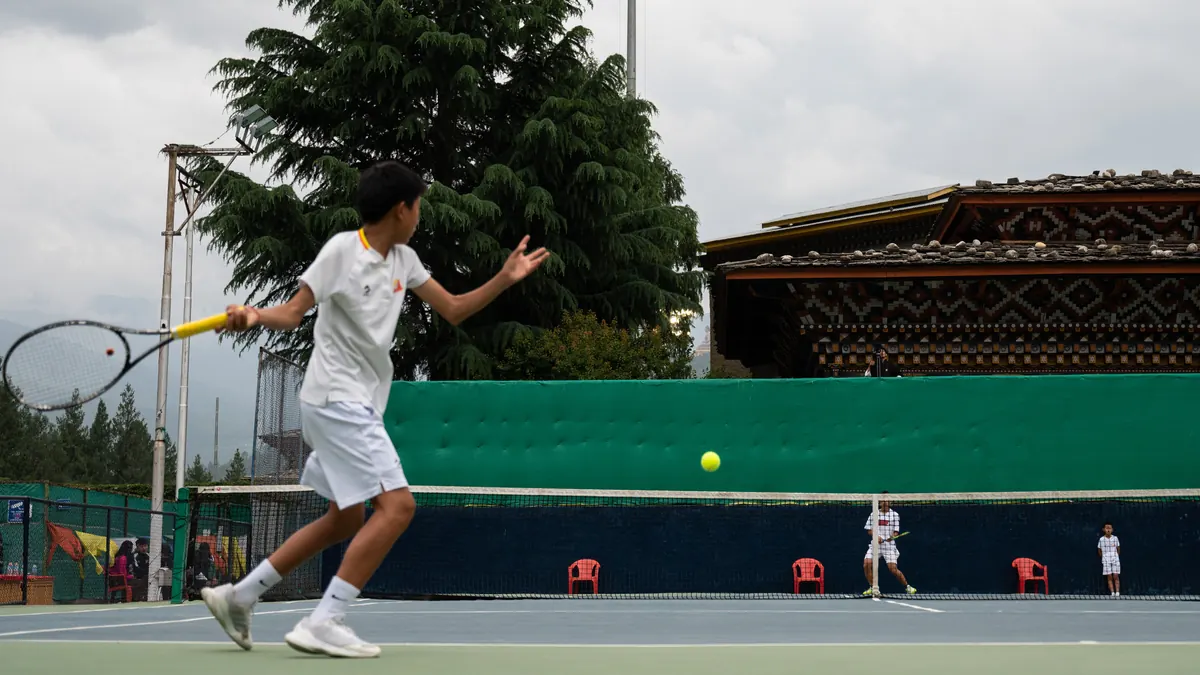 A junior hitting a forehand with the dzong roofline and the hills behind