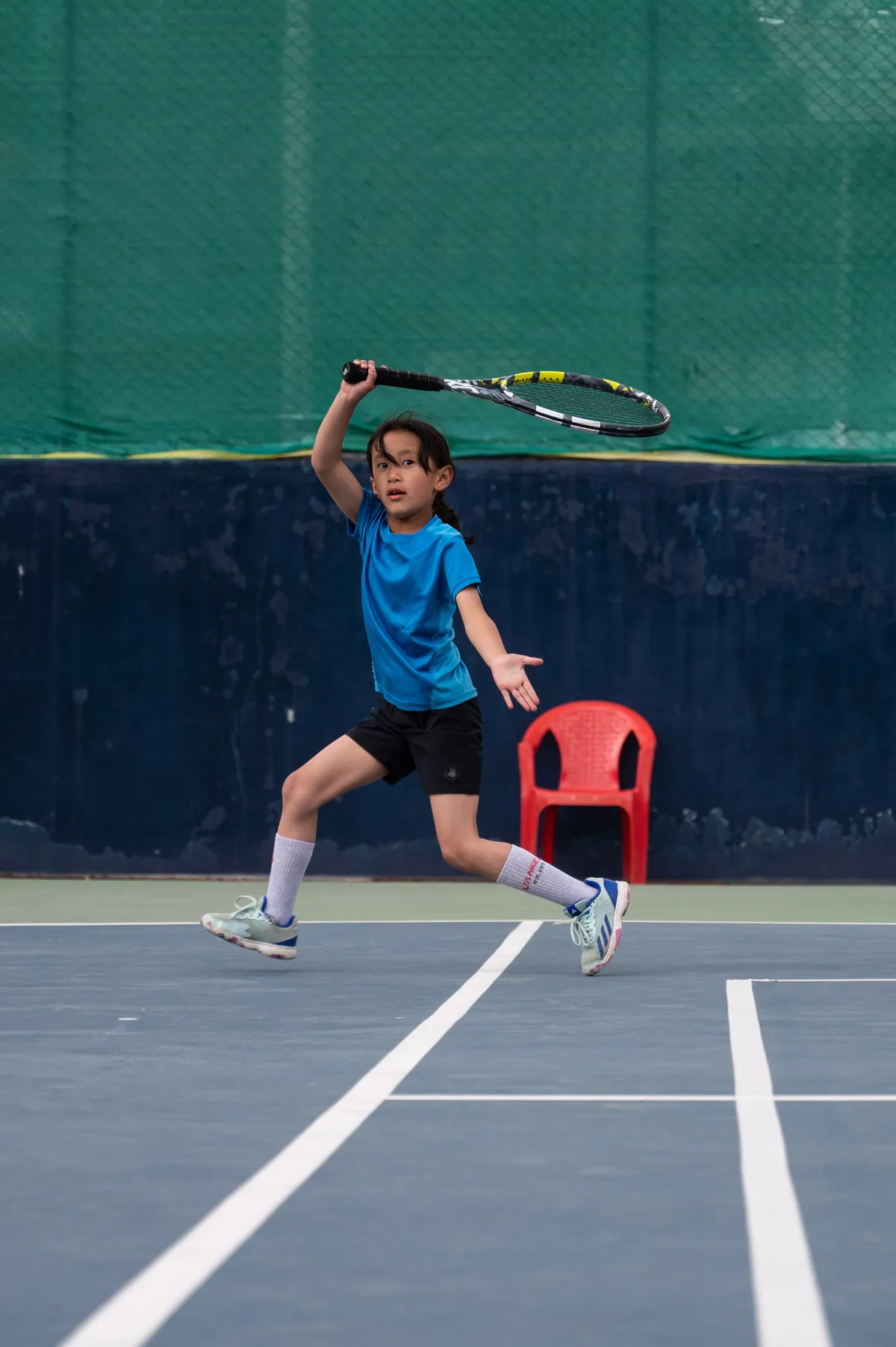 A junior in a blue shirt finishing a forehand at the National Tennis Centre