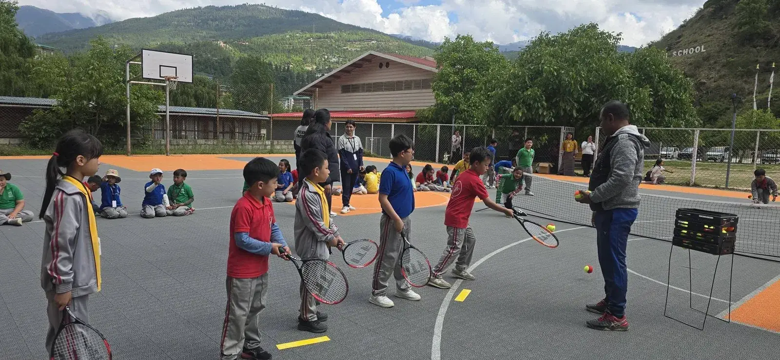 A coach running a tennis session for schoolchildren on an outdoor court in a Bhutanese valley