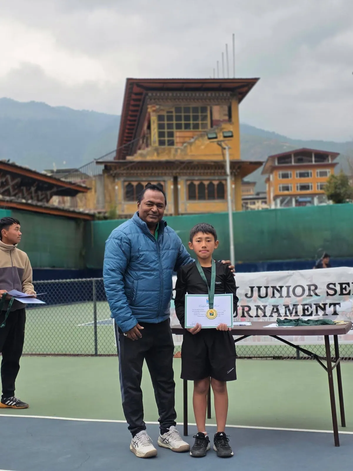 A coach standing with a junior champion holding his medal and certificate at a Junior Series presentation