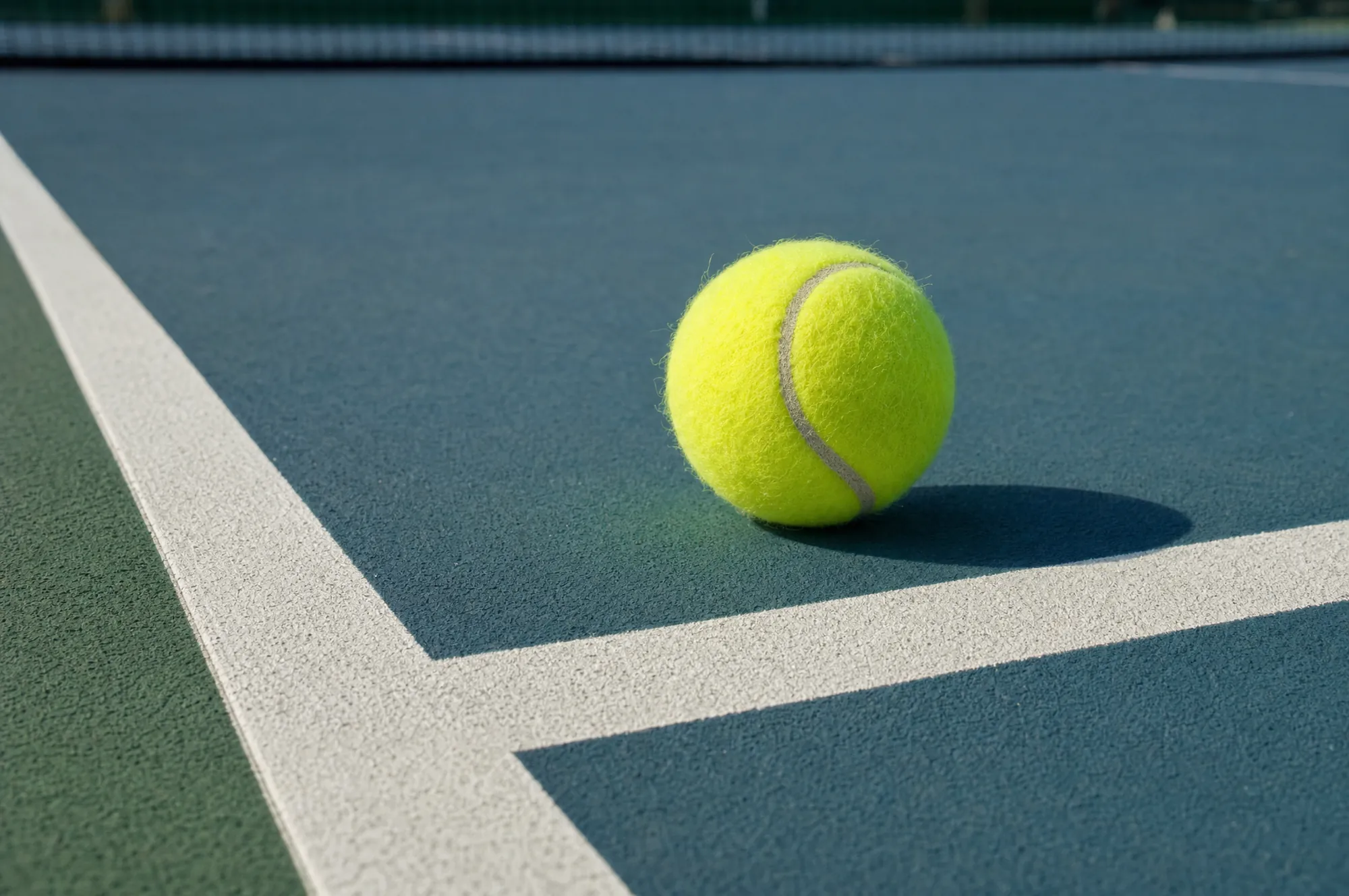 A tennis ball resting on the service line of a hard court, its shadow long in the afternoon sun
