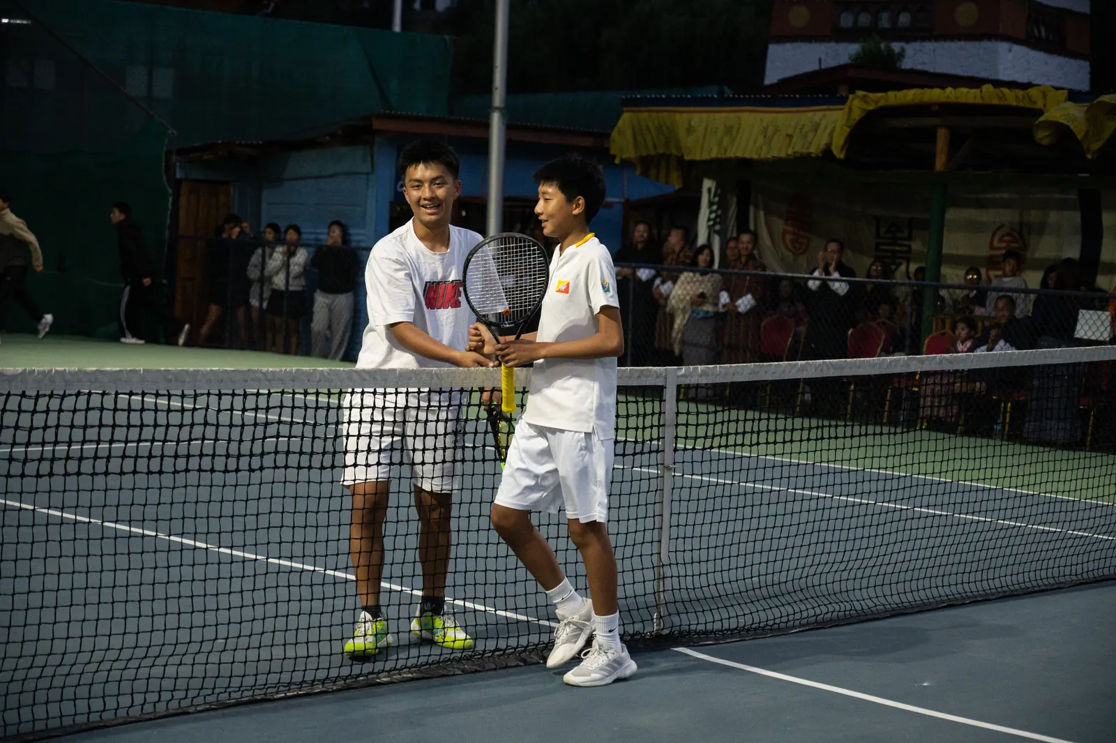 Two players shaking hands over the net at the end of a match, spectators behind