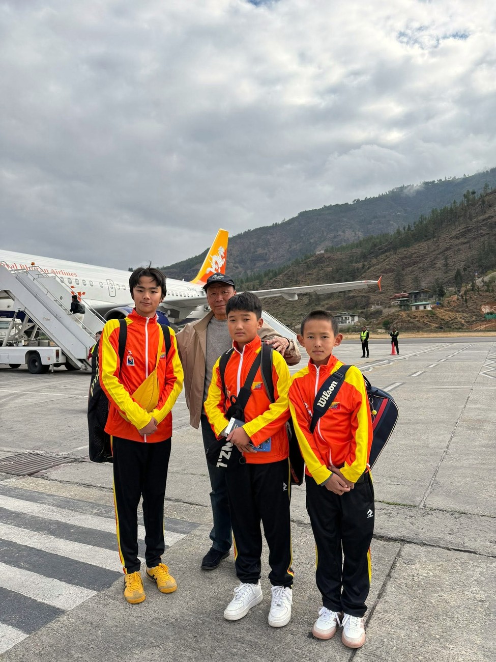Three junior players in national team tracksuits with their coach on the tarmac at Paro, a Drukair aircraft behind them