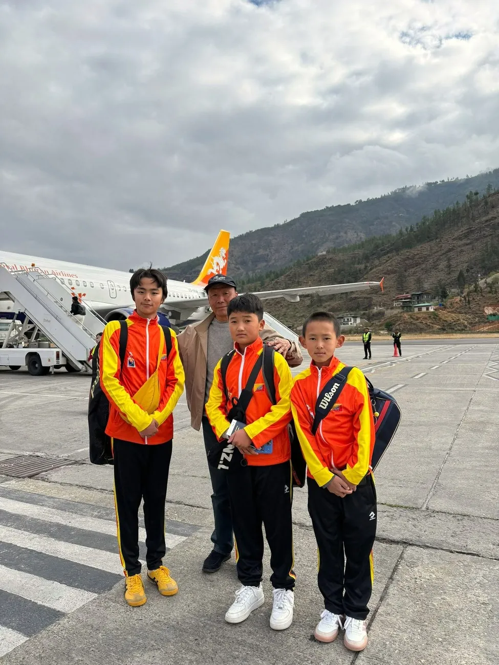 Three junior players in national team tracksuits with their coach on the tarmac at Paro, a Drukair aircraft behind them